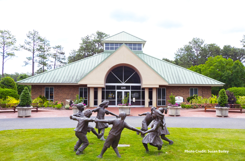 The Ball Gardens Visitor Center building is shown from the front, with landscaped planters and a sculpture of children holding hands on the lawn in the foreground.