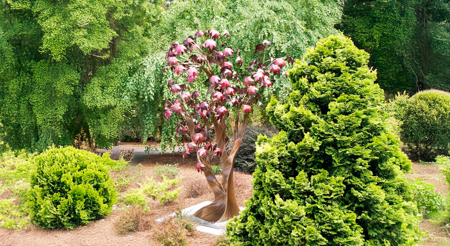 Metal tree sculpture with twisting trunk and pink leaf forms displayed among shrubs and pine straw in a landscaped garden setting.