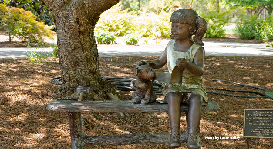 Bronze sculpture of a young girl sitting on a bench beside a dog, set beneath a tree in a garden landscape.