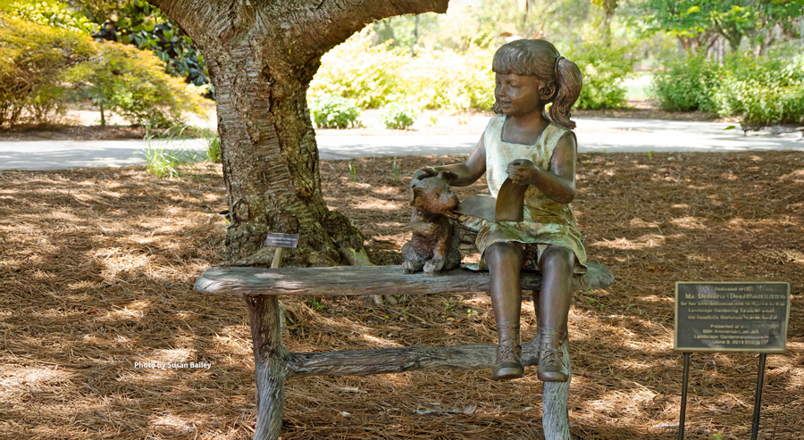 Bronze sculpture of a young girl sitting on a bench beside a dog, set beneath a tree in a garden landscape.