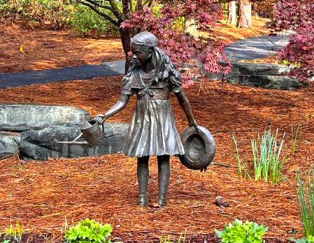 Bronze sculpture of a young girl holding a watering can and a hat, standing in a garden setting with stone features and autumn foliage nearby.