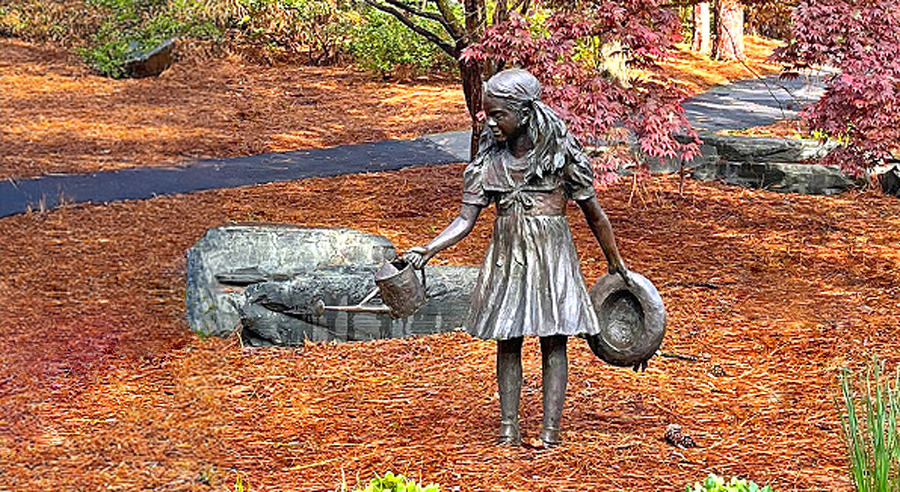 Bronze sculpture of a young girl holding a watering can and a hat, standing in a garden setting with stone features and autumn foliage nearby.