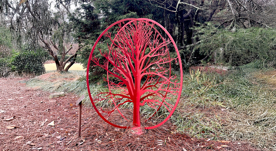 Bright red metal sculpture depicting the silhouette of an oak tree enclosed within a circular frame, installed among pine needles, ornamental grasses, and evergreen trees in the Sandhills Community College Horticultural Gardens.