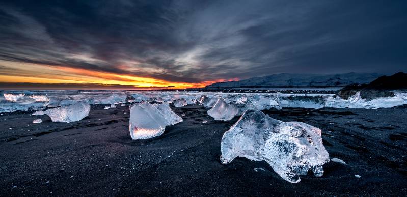 Chunks of clear ice rest on a black sand beach in Iceland as the sun sets along the horizon beneath dramatic, cloud-filled skies.