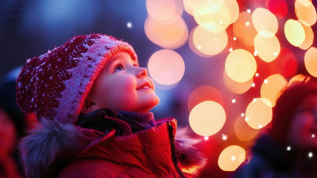 A smiling child in a winter hat looks up at glowing red and gold holiday lights.