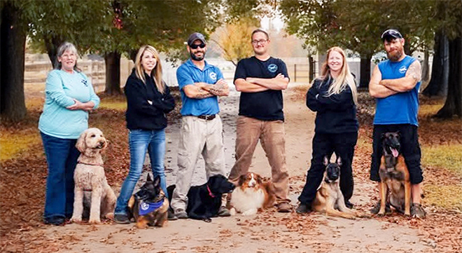 The Blue Cord K9 training team standing outdoors with several dogs on leashes along a tree-lined path.