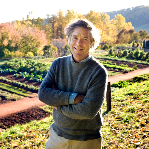 Peter Hatch stands with arms crossed in the Monticello vegetable garden, smiling amid cultivated rows of plants and autumn foliage.