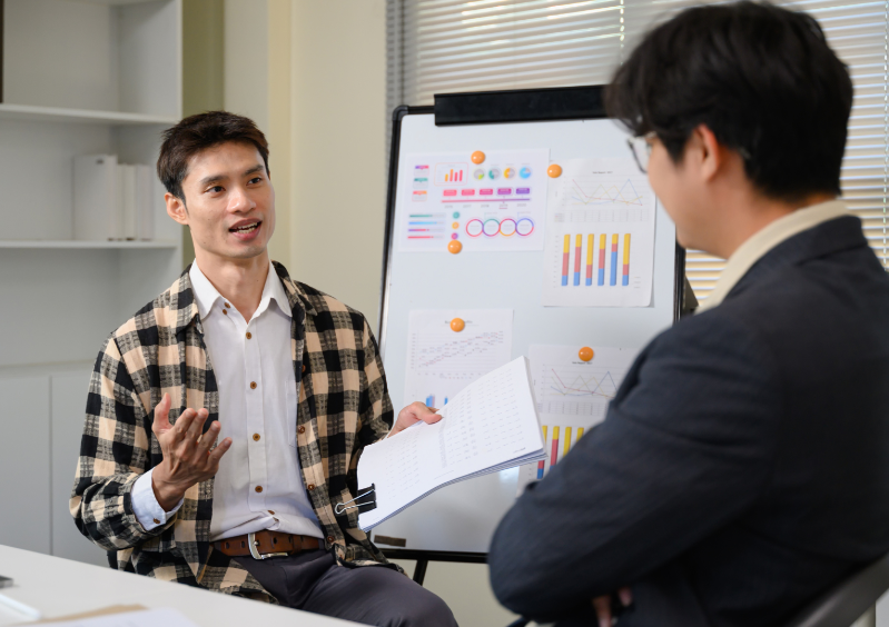 An instructor leads a one-on-one discussion with a participant in a classroom setting, using charts on a whiteboard to explain business concepts and data.