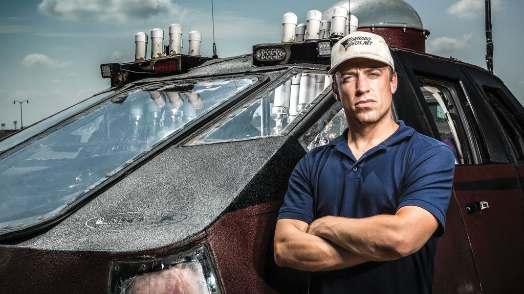Meteorologist and storm chaser Reed Timmer stands with his arms crossed in front of a storm-chasing vehicle.