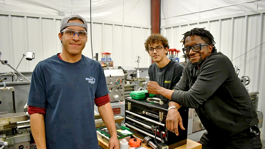 Three manufacturing students stand together in a machine shop with lathes and other machining equipment.