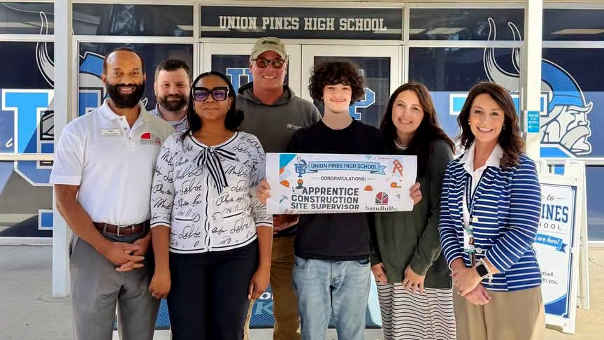 Union Pines High School student Connor Donathan holds a Construction Site Supervisor pre-apprenticeship sign while posing with six school and community partners outside Union Pines High School.