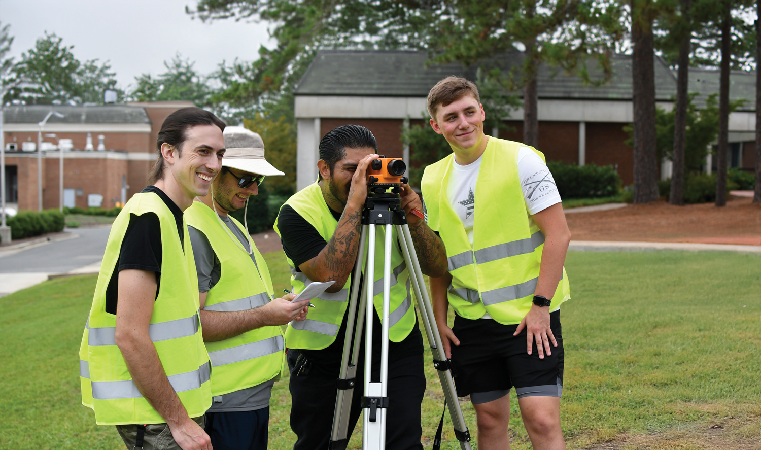 Surveying Students Group of students wearing reflective safety vests who are working together with surveying equipment during an outdoor training activity.